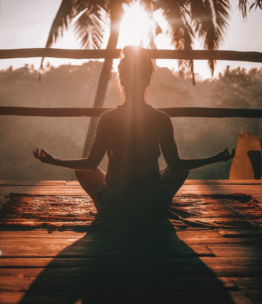 En kvinna sitter i en meditationsposition med slutna ögon och en lugn utstrålning, vilket ger intrycket av inre frid och avslappning. Denna bild symboliserar det holistiska tillvägagångssättet inom skönhet, där fokus ligger på att främja balans och välbefinnande för både kropp och sinne.